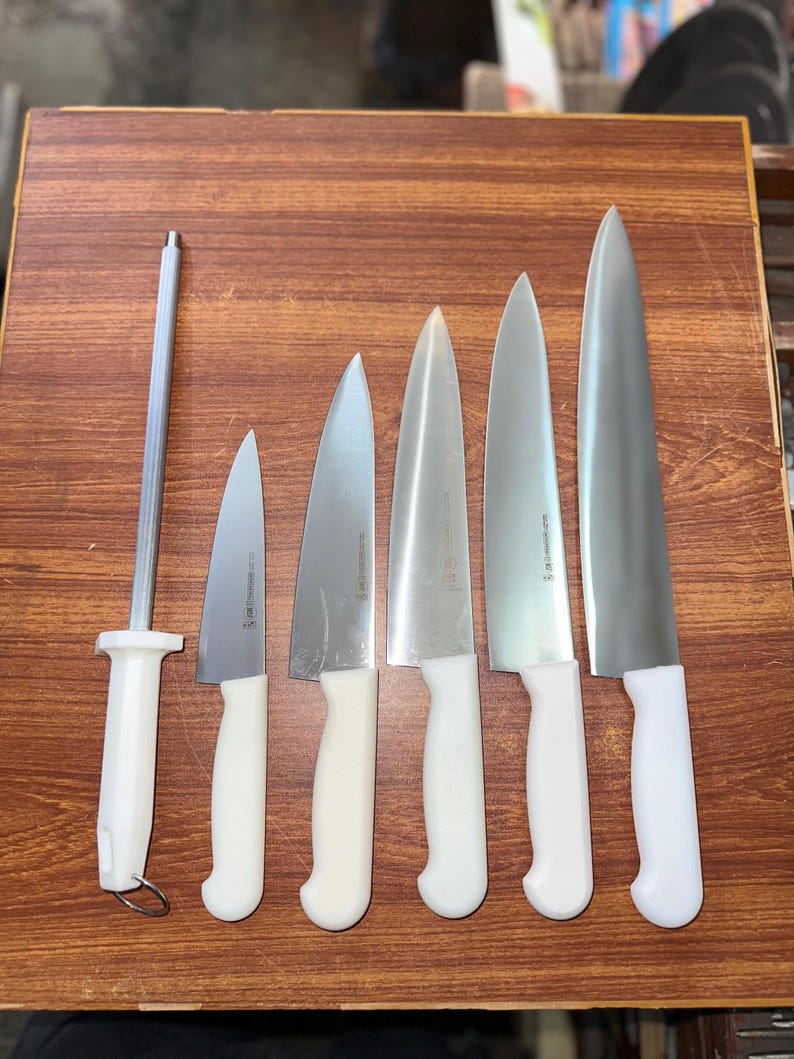 May include: A set of six stainless steel kitchen knives with white handles, ranging in size, and a sharpening steel. The knives are arranged on a wooden surface, showcasing their blades and handles.