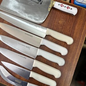 May include: A collection of stainless steel kitchen knives with white handles, including a large cleaver and several smaller blades. The cleaver has a brass bolster and a white handle with red and black accents. The knives are arranged on a wooden surface.