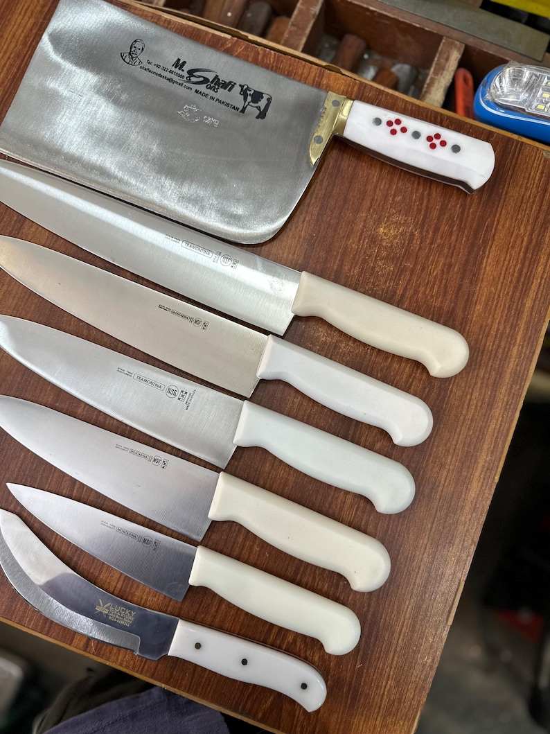 May include: A collection of stainless steel kitchen knives with white handles, including a large cleaver and several smaller blades. The cleaver has a brass bolster and a white handle with red and black accents. The knives are arranged on a wooden surface.