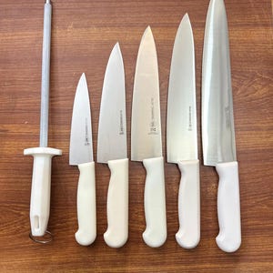 May include: A set of kitchen knives and a sharpening steel arranged on a wooden surface. The knives have silver blades and white handles. The sharpening steel has a white handle and a silver rod. The knives vary in size.