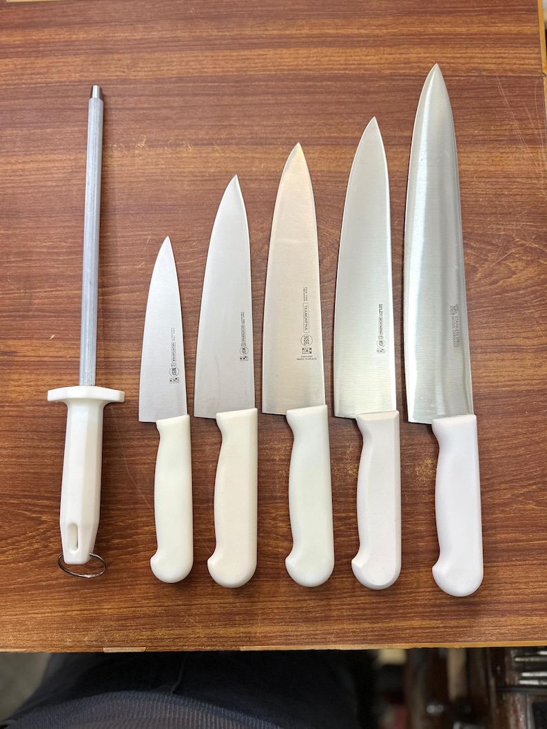 May include: A set of kitchen knives and a sharpening steel arranged on a wooden surface. The knives have silver blades and white handles. The sharpening steel has a white handle and a silver rod. The knives vary in size.