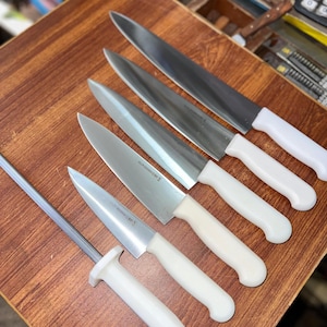 May include: A collection of stainless steel kitchen knives with white handles, arranged on a wooden surface. The set includes various sizes of chef's knives and a sharpening steel. The blades are sharp and ready for use.