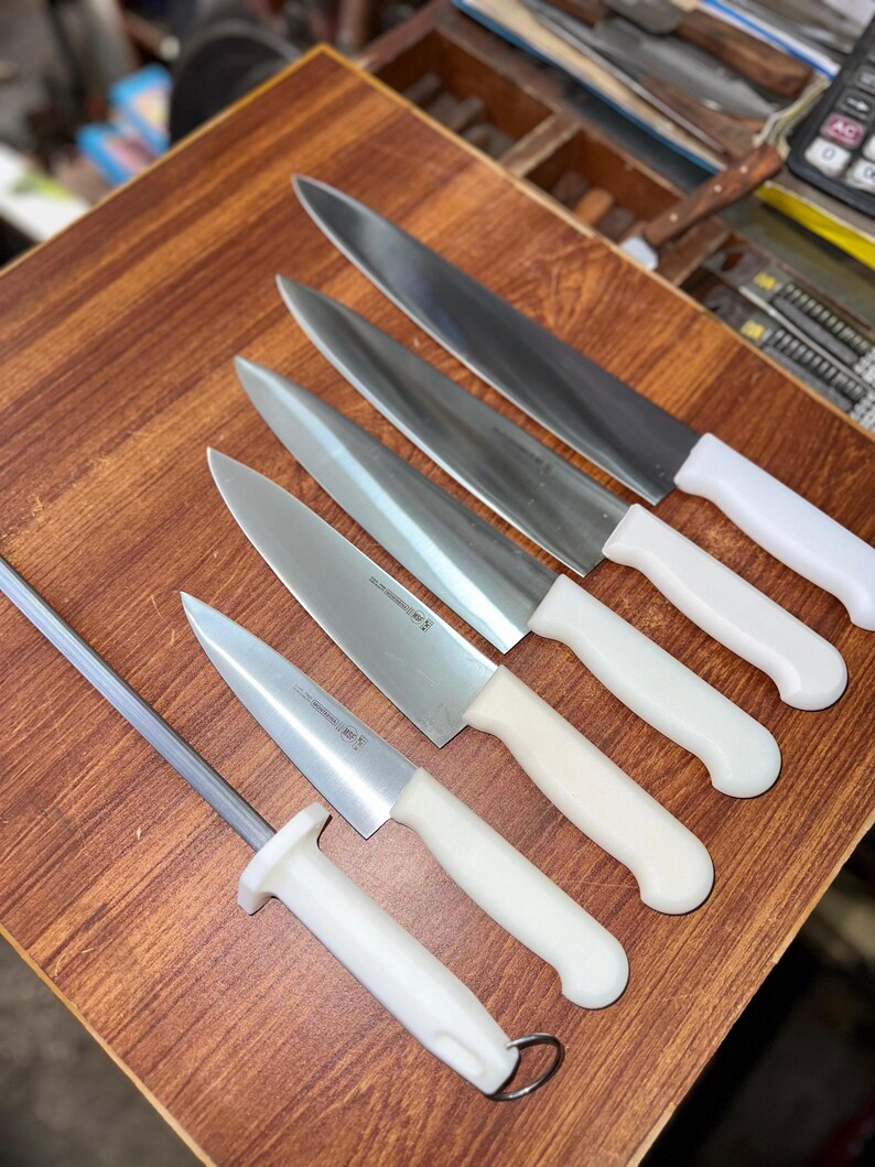 May include: A collection of stainless steel kitchen knives with white handles, arranged on a wooden surface. The set includes various sizes of chef's knives and a sharpening steel. The blades are sharp and ready for use.
