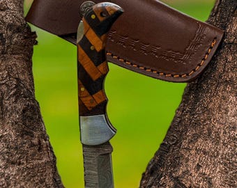 Op de afbeelding: Een vouwmes met een patroon houten handvat in bruin- en zwarttinten, en een zilveren lemmet. Het mes wordt gecombineerd met een bruine leren schede. De achtergrond is een vage groene en bruine kleur.