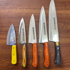 Op de afbeelding: Een assortiment van vijf keukenmessen met zilveren bladen en houten handvatten. De handvatten zijn in verschillende kleuren, waaronder geel, oranje en zwart. De bladen hebben de tekst "LUCKY TOKA STORE" erop.