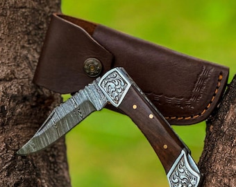 Könnte beinhalten: Ein Klappmesser mit gemusterter Klinge und dunklem Holzgriff. Der Griff hat dekorative Metallakzente. Eine braune Lederscheide mit Druckknopfverschluss befindet sich hinter dem Messer. Das Messer liegt auf einem Ast.