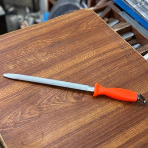 May include: A silver knife sharpening steel with an orange handle rests on a wooden surface. The steel is straight and has a pointed tip. The handle has a loop at the end.