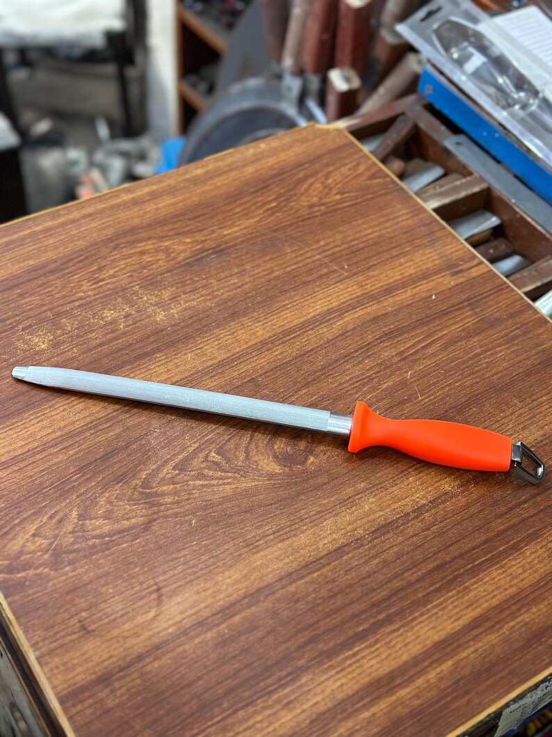 May include: A silver knife sharpening steel with an orange handle rests on a wooden surface. The steel is straight and has a pointed tip. The handle has a loop at the end.