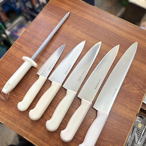 May include: A set of kitchen knives and a sharpening steel arranged on a wooden surface. The knives have silver blades and off-white handles. The sharpening steel has a silver rod and an off-white handle. The knives vary in size.