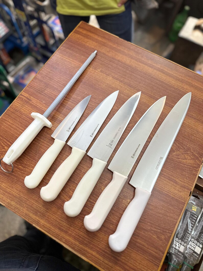 May include: A set of kitchen knives and a sharpening steel arranged on a wooden surface. The knives have silver blades and off-white handles. The sharpening steel has a silver rod and an off-white handle. The knives vary in size.