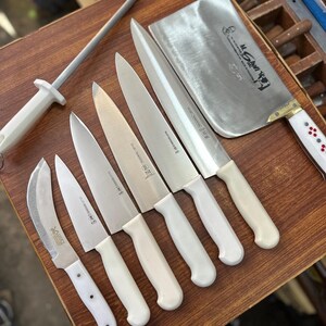 May include: A collection of stainless steel kitchen knives with white handles, including a cleaver, chef's knives, and a paring knife, arranged on a wooden surface. A sharpening steel is also present. The knives are designed for professional or home use.