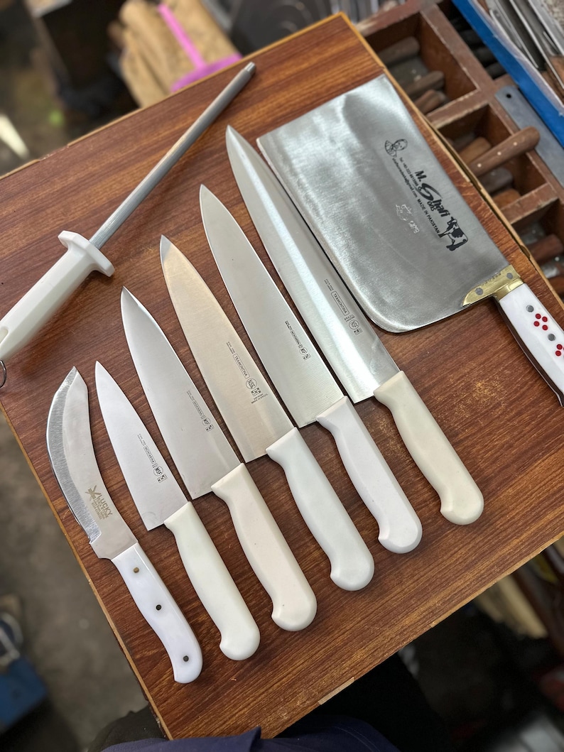 May include: A collection of stainless steel kitchen knives with white handles, including a cleaver, chef's knives, and a paring knife, arranged on a wooden surface. A sharpening steel is also present. The knives are designed for professional or home use.