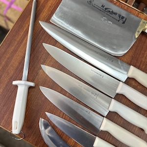 May include: A collection of stainless steel kitchen knives with white handles, including a cleaver, chef's knives, and a sharpening steel. The blades are arranged on a wooden surface, showcasing their sharp edges and professional design.