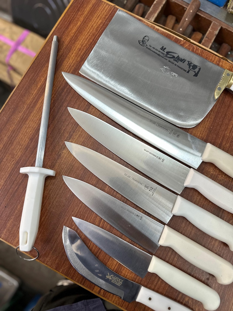 May include: A collection of stainless steel kitchen knives with white handles, including a cleaver, chef's knives, and a sharpening steel. The blades are arranged on a wooden surface, showcasing their sharp edges and professional design.