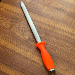 May include: A knife sharpening steel with a textured, silver-colored rod and a bright orange handle. The handle has a curved ergonomic design and a metal loop at the base. The steel is designed for sharpening kitchen knives.