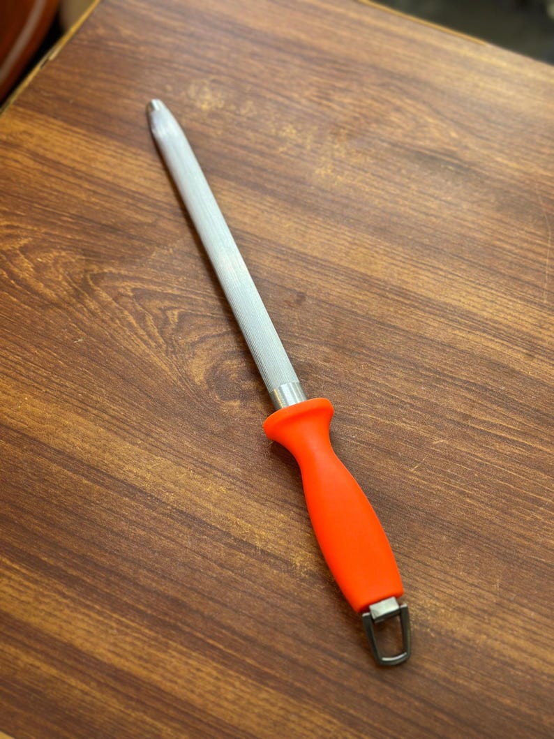 May include: A knife sharpening steel with a textured, silver-colored rod and a bright orange handle. The handle has a curved ergonomic design and a metal loop at the base. The steel is designed for sharpening kitchen knives.
