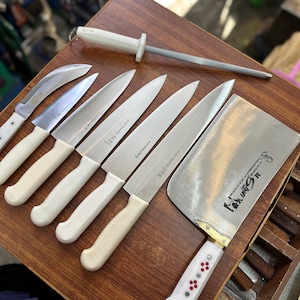 May include: A collection of stainless steel kitchen knives with white handles and a sharpening steel. The knives include a cleaver, paring knives, and chef's knives, arranged on a wooden surface. The cleaver has a white handle with red and white accents.