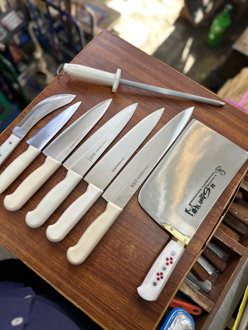 May include: A collection of stainless steel kitchen knives with white handles and a sharpening steel. The knives include a cleaver, paring knives, and chef's knives, arranged on a wooden surface. The cleaver has a white handle with red and white accents.