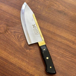 Könnte beinhalten: Ein Edelstahl-Kochmesser mit gebogener Klinge und schwarzem Griff. Der Griff ist mit Messing und goldenen Nieten verziert. Die Klinge trägt den Text "LUCKY" und andere Texte.