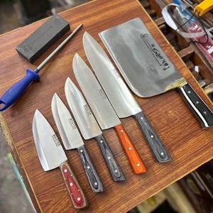 Może przedstawiać: Zestaw noży kuchennych i tasak z drewnianymi uchwytami w różnych kolorach, w tym czerwonym, pomarańczowym i szarym. Obecny jest również pręt do ostrzenia i kamień, wszystko ułożone na drewnianej powierzchni.