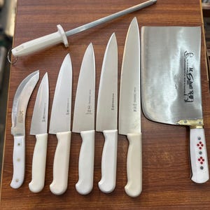 May include: A collection of kitchen knives and a sharpening steel arranged on a wooden surface. The knives have white handles and silver blades. A large cleaver with a white handle and red accents is also present. The knives vary in size and shape.