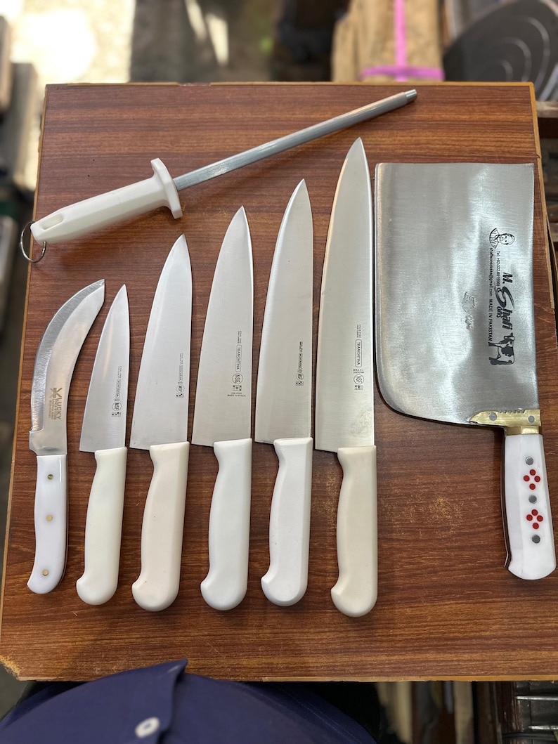 May include: A collection of kitchen knives and a sharpening steel arranged on a wooden surface. The knives have white handles and silver blades. A large cleaver with a white handle and red accents is also present. The knives vary in size and shape.