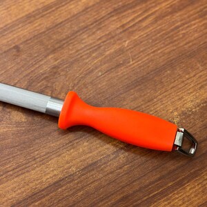May include: A knife sharpening steel with a bright orange handle and a silver steel rod. The handle has a curved design for a comfortable grip. The steel has a metal loop at the end.