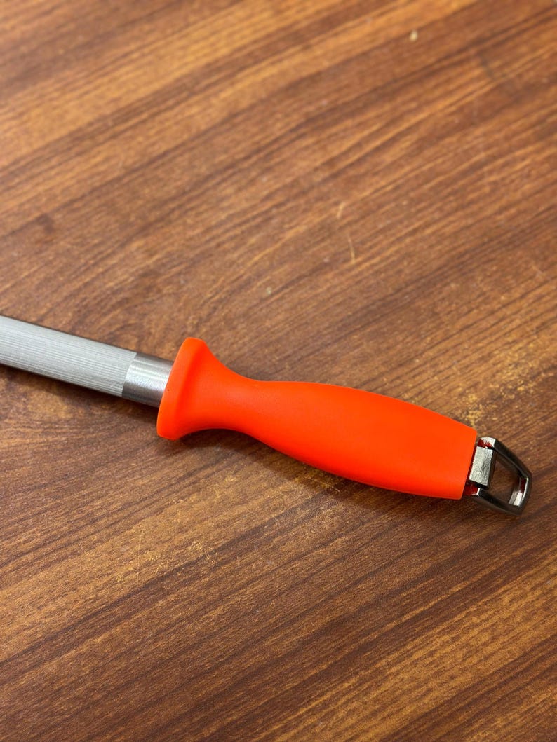 May include: A knife sharpening steel with a bright orange handle and a silver steel rod. The handle has a curved design for a comfortable grip. The steel has a metal loop at the end.