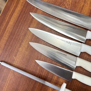 May include: A set of stainless steel kitchen knives with white handles and a sharpening steel. The knives are arranged on a wooden surface, showcasing their sharp blades. The set includes various sizes for different culinary tasks.
