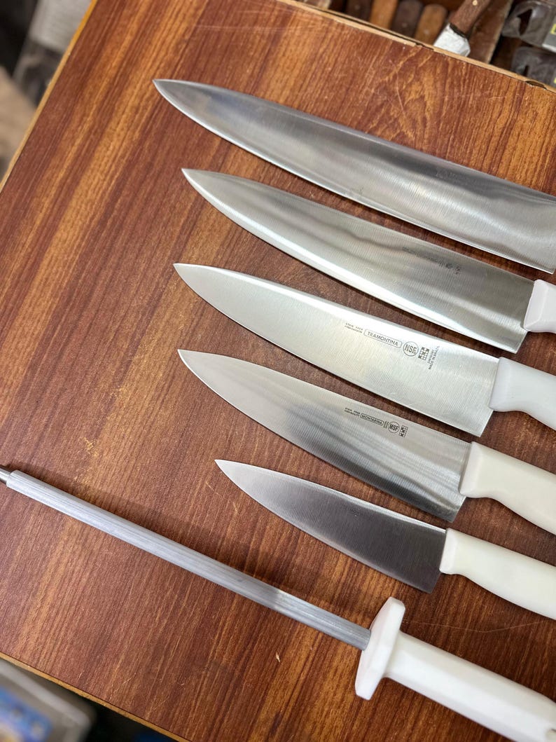 May include: A set of stainless steel kitchen knives with white handles and a sharpening steel. The knives are arranged on a wooden surface, showcasing their sharp blades. The set includes various sizes for different culinary tasks.