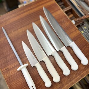 May include: A collection of kitchen knives and a sharpening steel arranged on a wooden surface. The knives have silver blades and white handles. The sharpening steel has a silver rod and a white handle. The knives vary in size.