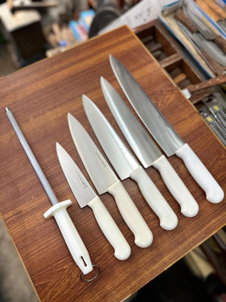 May include: A collection of kitchen knives and a sharpening steel arranged on a wooden surface. The knives have silver blades and white handles. The sharpening steel has a silver rod and a white handle. The knives vary in size.