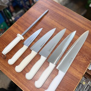 May include: A set of kitchen knives and a sharpening steel arranged on a wooden surface. The knives have silver blades and white handles. The sharpening steel has a gray rod and a white handle.