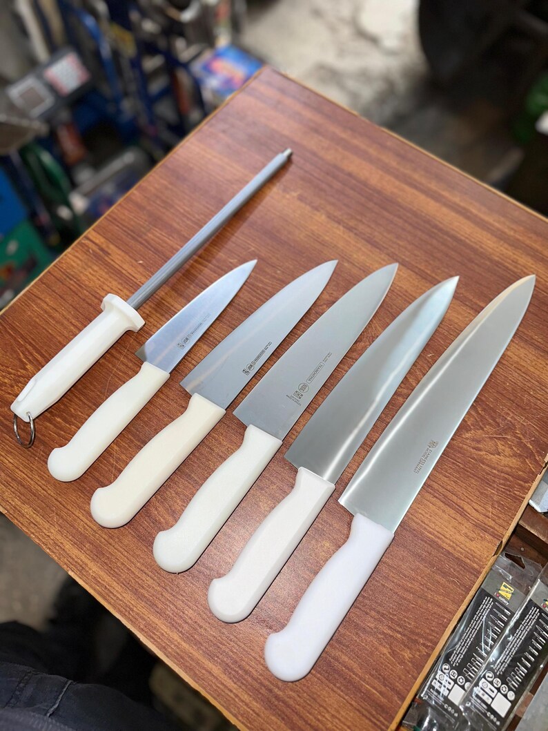 May include: A set of kitchen knives and a sharpening steel arranged on a wooden surface. The knives have silver blades and white handles. The sharpening steel has a gray rod and a white handle.
