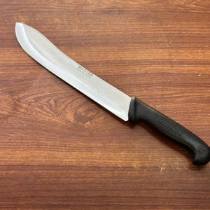 Puede incluir: Un cuchillo de acero inoxidable con mango negro. La hoja es plateada y tiene la palabra "MASTER" impresa. El mango tiene la palabra "SAMPRENE" impresa. El cuchillo está sobre una superficie de madera.