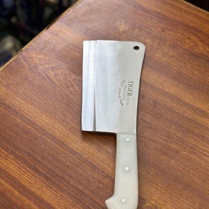 May include: A stainless steel cleaver with a white handle. The blade is rectangular with a curved handle, and the text "TIGER SPECIAL Meat Cleaver High Carbon-no Stain" is printed on the blade. The cleaver is on a wooden surface.