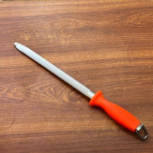May include: A knife sharpening steel with a textured, silver-colored rod and a bright orange handle. The handle has a loop at the end for hanging. The tool is designed for sharpening kitchen knives.
