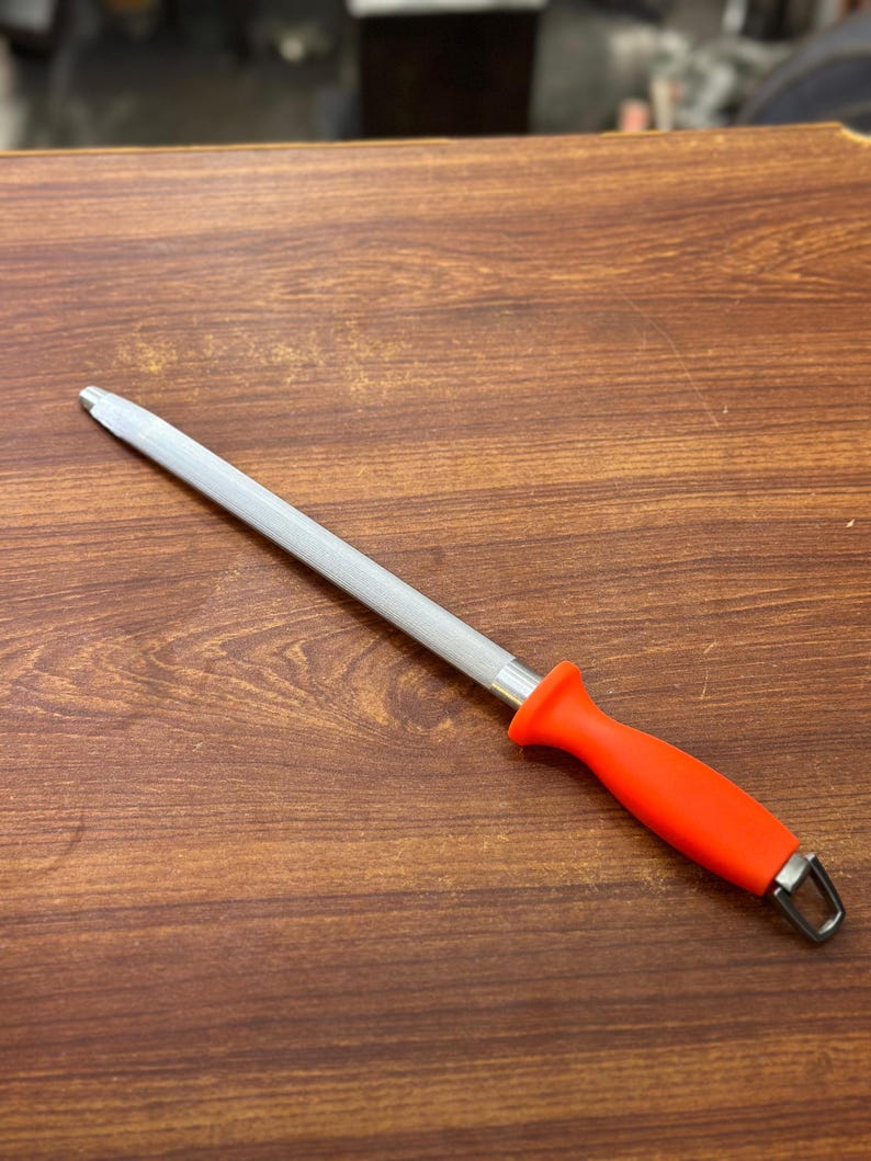 May include: A knife sharpening steel with a textured, silver-colored rod and a bright orange handle. The handle has a loop at the end for hanging. The tool is designed for sharpening kitchen knives.