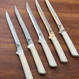 May include: Five stainless steel fillet knives with off-white handles are arranged diagonally on a wooden surface. Each knife has a long, slender blade and a bolster where the blade meets the handle. The handles are a light cream color.