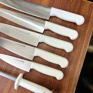May include: A collection of stainless steel kitchen knives with white handles, including a sharpening steel. The knives are arranged on a wooden surface, showcasing their blades and handle designs. The set includes various sizes and shapes for different culinary tasks.