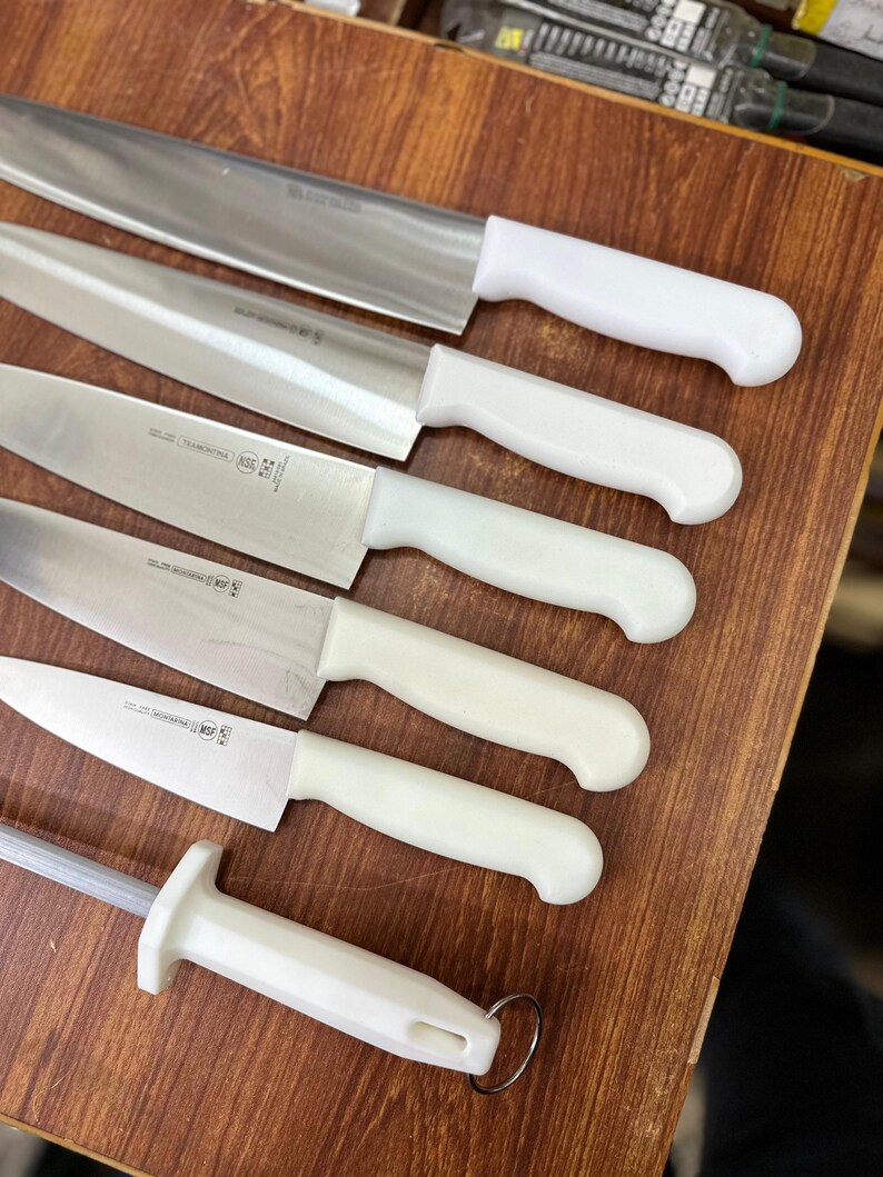 May include: A collection of stainless steel kitchen knives with white handles, including a sharpening steel. The knives are arranged on a wooden surface, showcasing their blades and handle designs. The set includes various sizes and shapes for different culinary tasks.