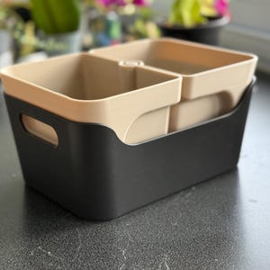 Peut inclure: Un bac de rangement bicolore avec un extérieur noir et des compartiments intérieurs beiges. Le bac a une forme rectangulaire avec des coins arrondis et des poignées découpées. L'intérieur est divisé en sections, adapté pour organiser des articles. Le bac est en plastique.