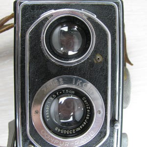 May include: A vintage Ikoflex twin-lens reflex camera with a black textured body and chrome accents. The front features two circular lenses, with the lower lens labeled Zeiss Ikon, Tessar 1:3.5 f=7.5cm, Carl Zeiss Jena Nr. 2338548, and Compur-Rapid.