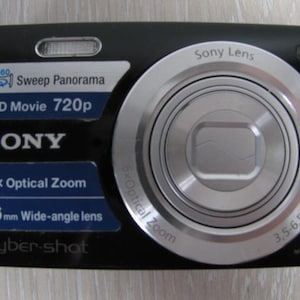 May include: A black Sony Cyber-shot digital camera featuring a silver lens with 6x optical zoom, 26mm wide-angle lens, 16.1 megapixels, and 720p HD movie capability. The front displays Sweep Panorama branding and a built-in flash unit.