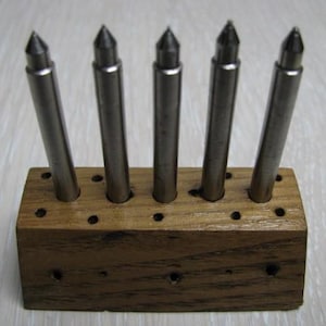 May include: Five cylindrical metal tools with pointed tips are inserted into a rectangular wooden block. The block features multiple pre-drilled holes arranged in two rows, providing a stable base for the upright steel implements.