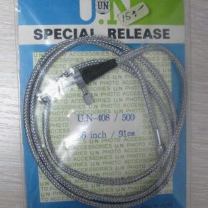 Puede incluir: Un cable disparador de cámara trenzado plateado, modelo U.N-408 / 500, que mide 91 cm de largo. El cable está empaquetado en una bolsa de plástico transparente con una tarjeta de respaldo azul y verde con el texto U.N SPECIAL RELEASE y U.N COMPANY LTD.