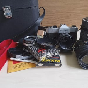 May include: A collection of vintage camera equipment, including a Yashica camera, lenses, filters, and a black leather camera bag. The lenses and filters are various sizes and brands. A red camera strap is also visible.