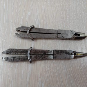 May include: Two vintage metal pliers with a weathered, dark gray finish. Each tool features a long, slender design with a sliding metal ring mechanism near the handle end. The metal surfaces show signs of oxidation and wear, with visible texture.