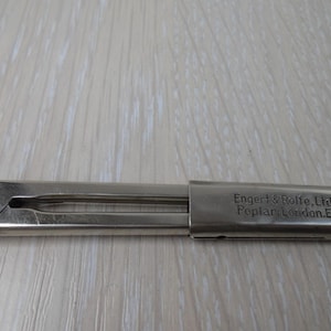 Peut inclure: Un outil pliant en métal argenté avec une extrémité arrondie et un point de pivot sur le côté opposé. La surface présente le texte gravé Engert & Rolfe, Ltd. Poplar, London, E. L'objet a une finition métallique lisse et une forme allongée.