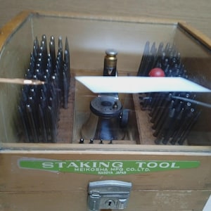May include: A wooden watchmaker's staking tool set housed in a box with a curved glass lid. The interior contains numerous metal punches and a central staking base. A green label on the front reads STAKING TOOL MEIKOSHA MFG CO. LTD. NAGOYA JAPAN.